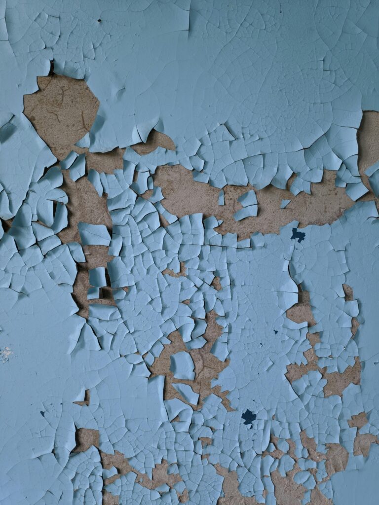 Close-up of a weathered wall with cracking and peeling blue paint, revealing rough texture beneath.